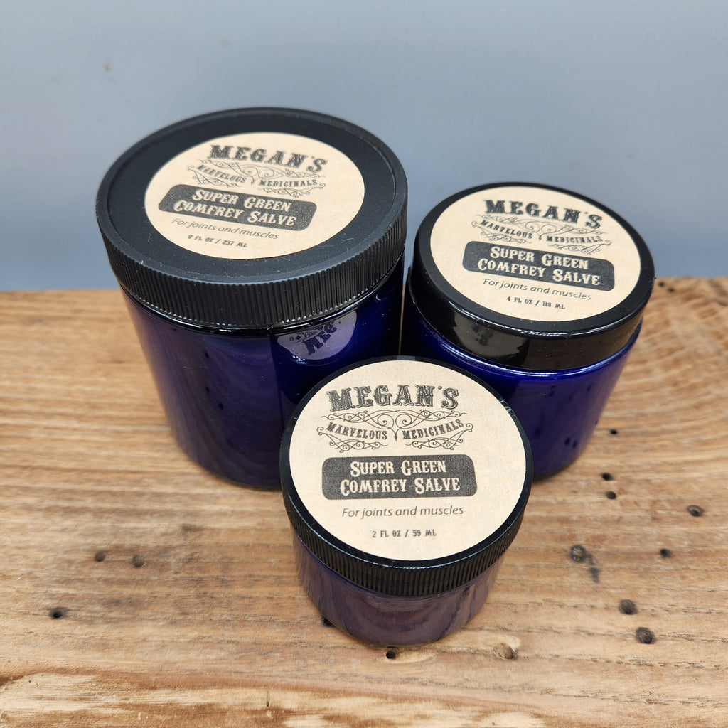 Three jars of Megan's Super Green Comfort Salve on a wooden surface with a light blue background.