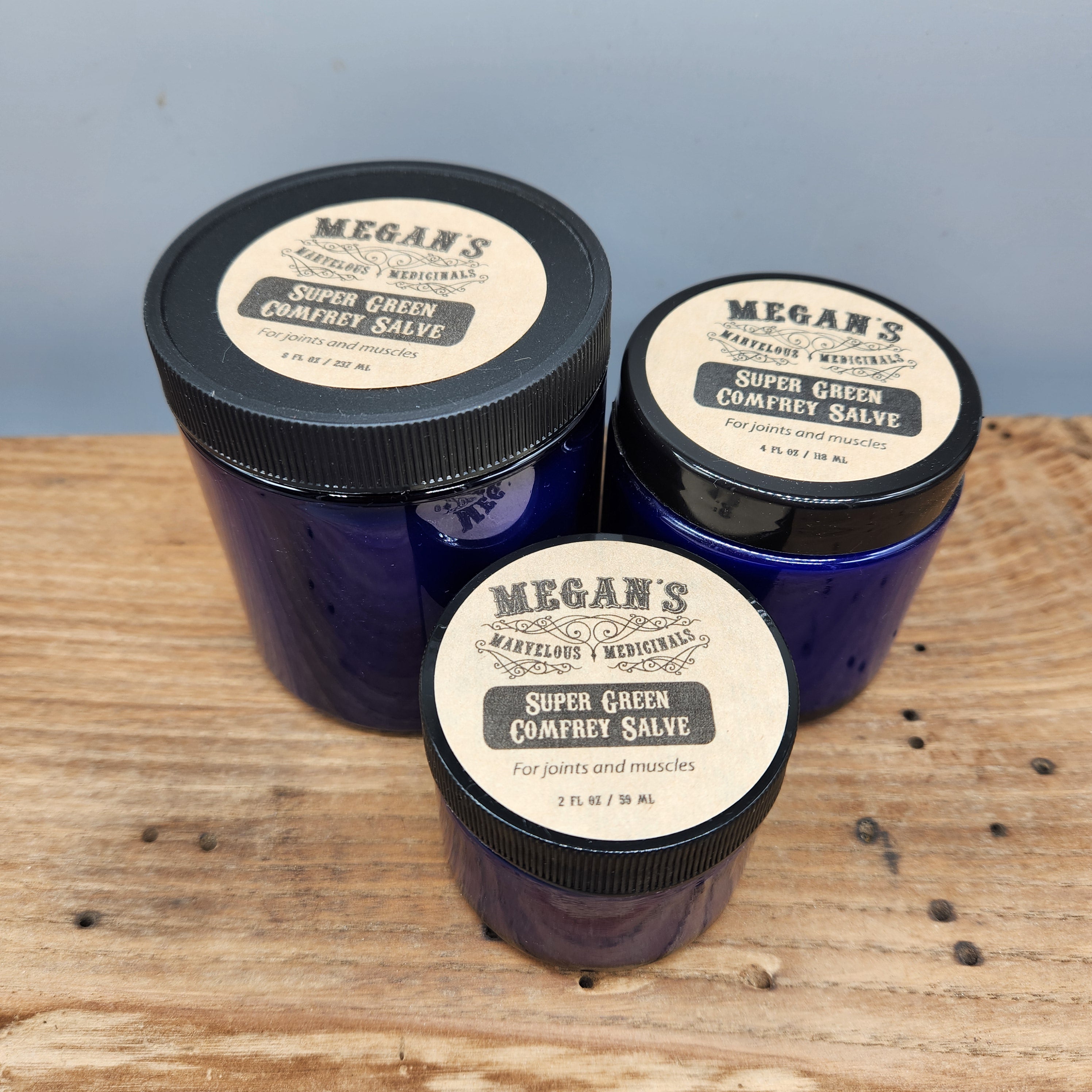 Three jars of Megan's Super Green Comfort Salve on a wooden surface with a light blue background.