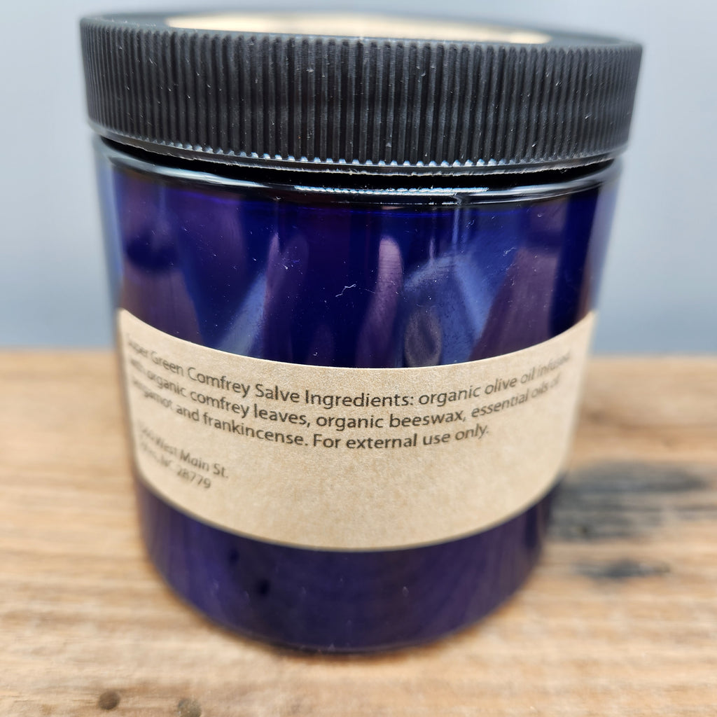 Blue jar with a black lid on a wooden surface. showing ingredients label for Megan's Marvelous Medicinals super green comfrey salve.