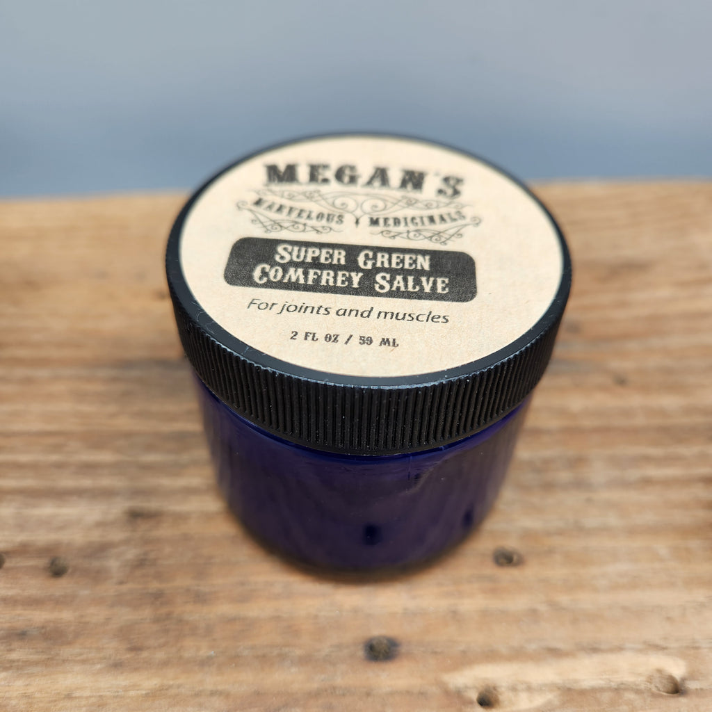 Blue jar of Megan's Marvelous Medicinals 2 oz super green comfrey salve on a wooden surface with a blurred blue background.