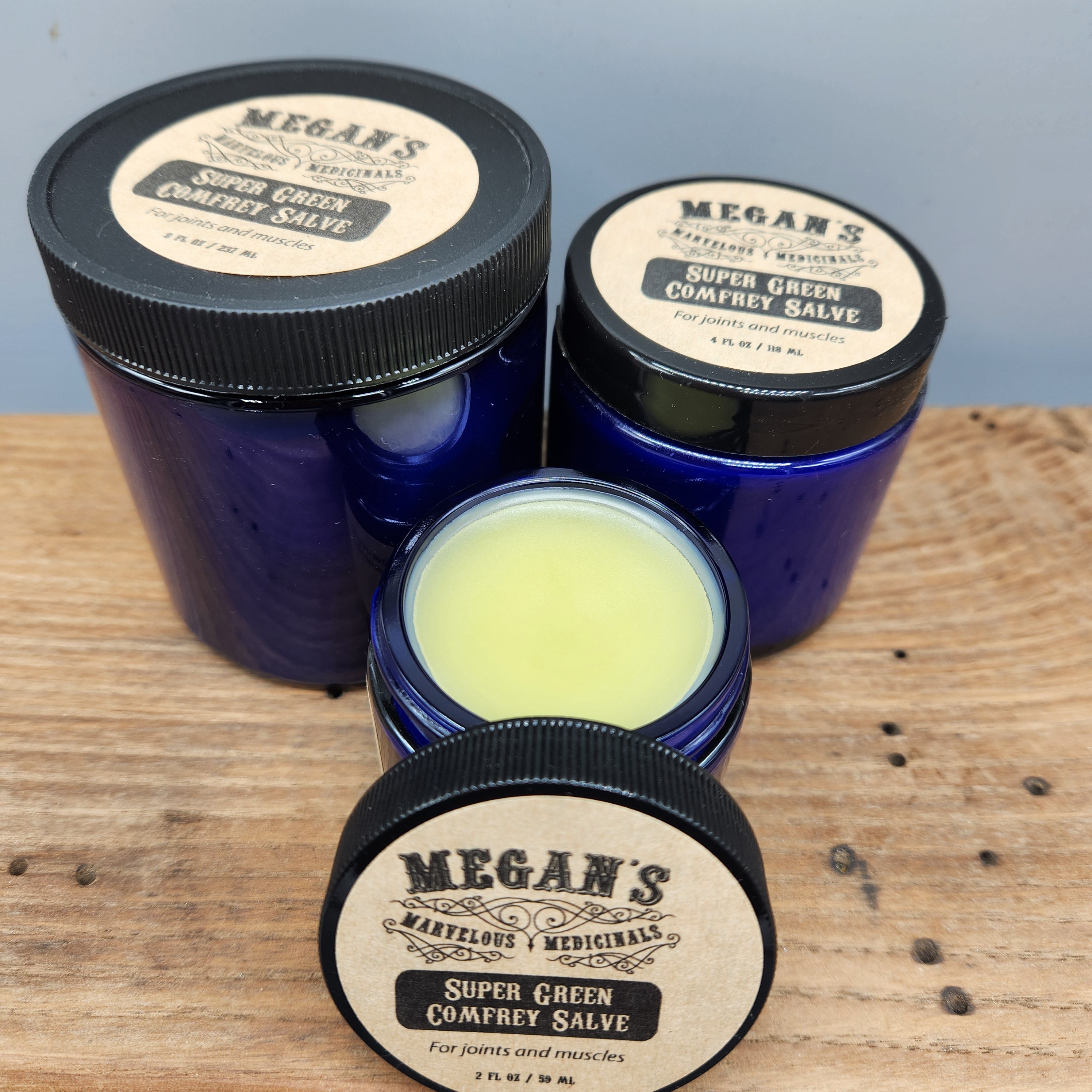 Three blue jars with black lids on a wooden surface filled with Megan's Marvelous Medicinals Super Green Comfrey salve
